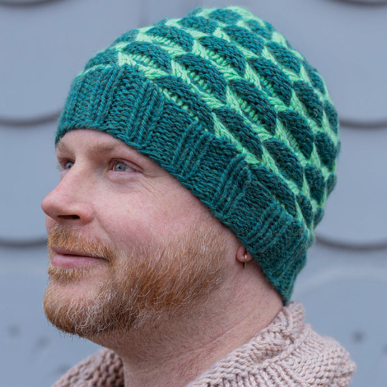This image shows designer Stephen West wearing a beautifully knitted hat in two shades of green.
