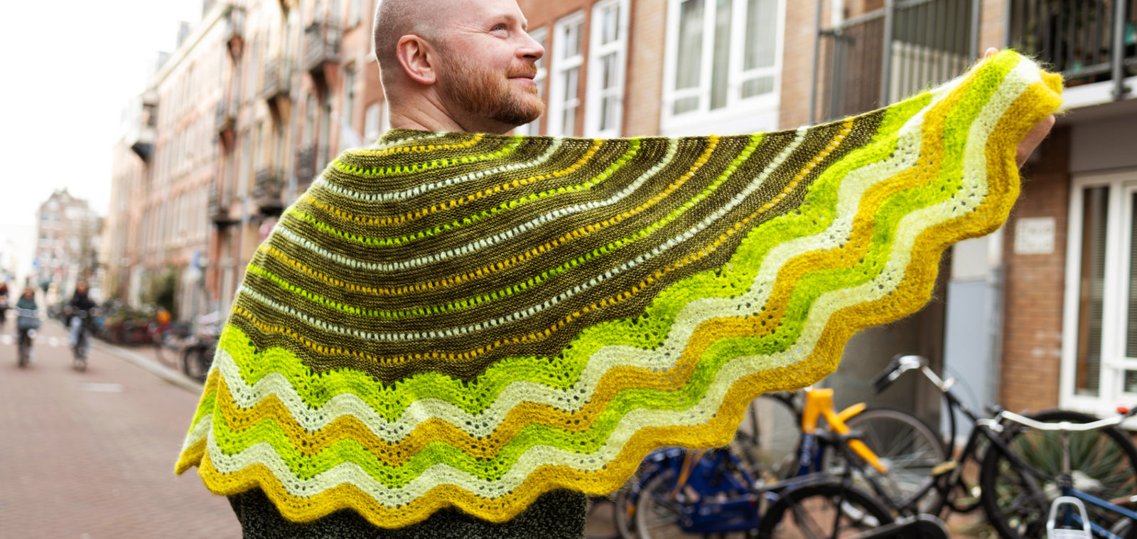 This image shows Stephen West wearing a  beautifully knitted sample of the Suri Shimmy Shawl in green and yellow gradients. The background shows a street in Amsterdam.