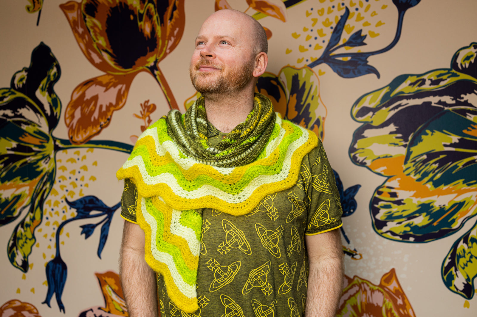 This image shows Stephen West wearing a  beautifully knitted sample of the Suri Shimmy Shawl around his neck in green and yellow gradients. The background shows a flower wallpaper. 