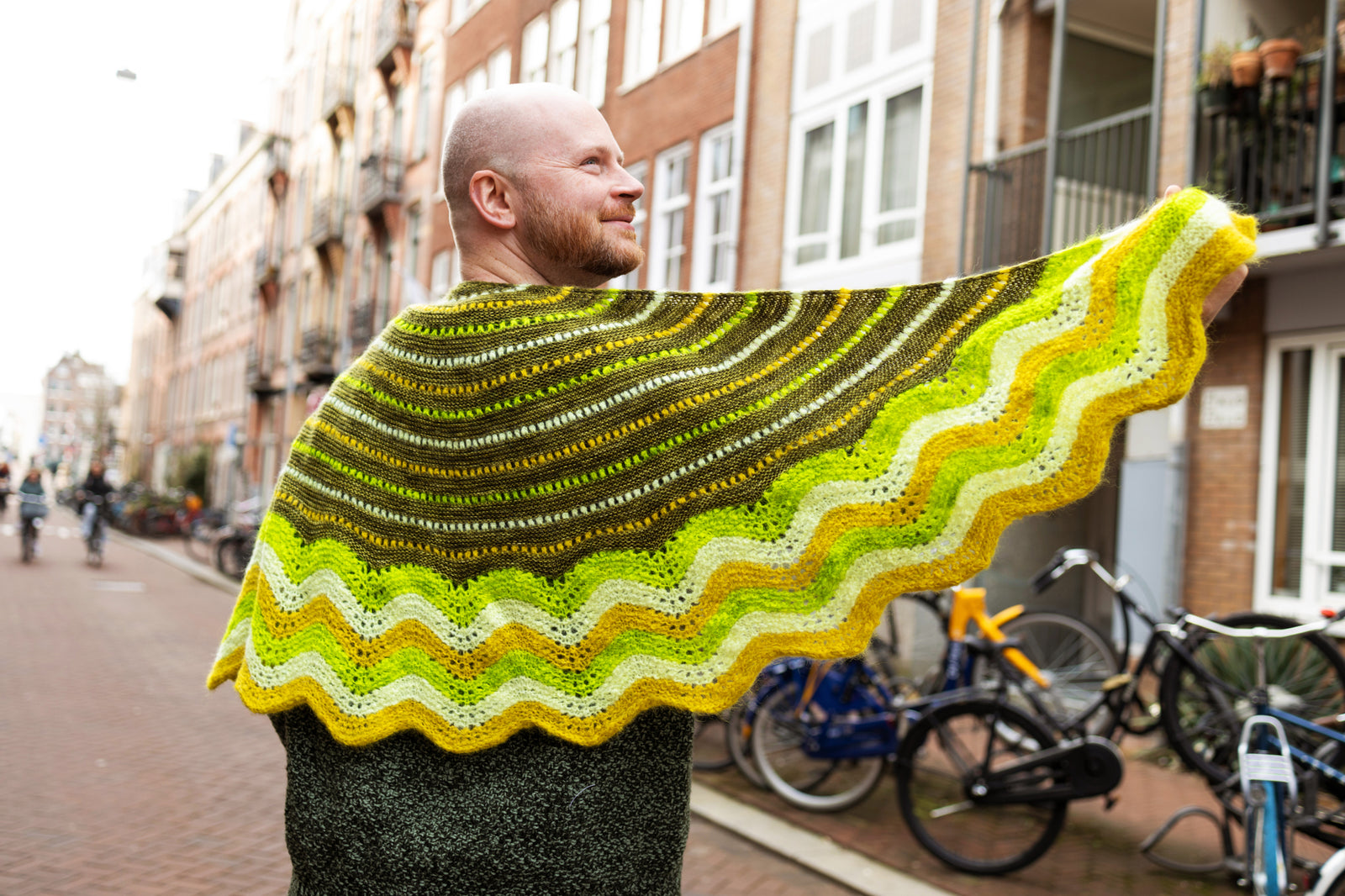 This image shows Stephen West wearing a  beautifully knitted sample of the Suri Shimmy Shawl in green and yellow gradients. The background shows a street in Amsterdam.