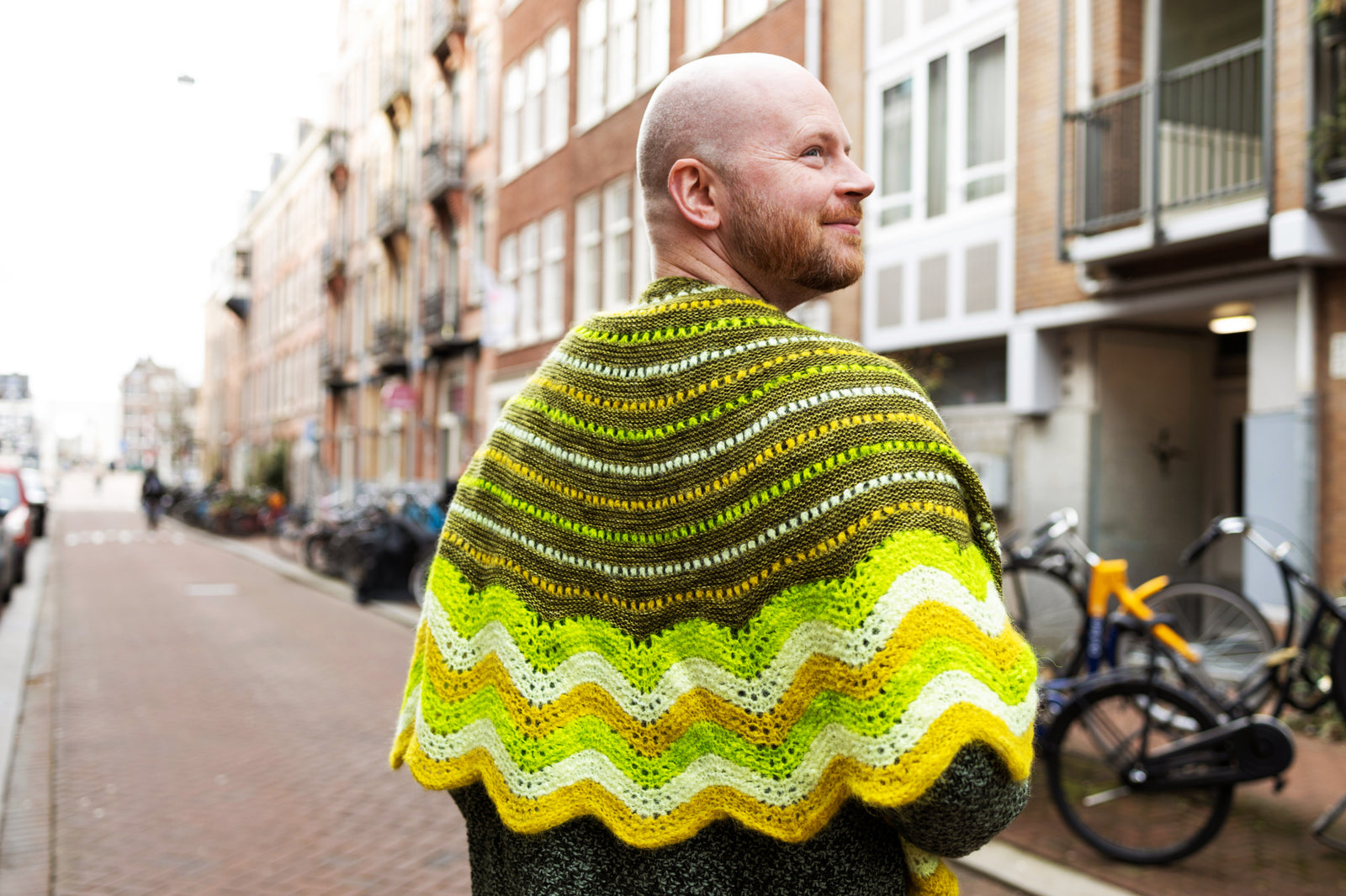 This image shows Stephen West wearing a  beautifully knitted sample of the Suri Shimmy Shawl in green and yellow gradients. The background shows a street in Amsterdam.