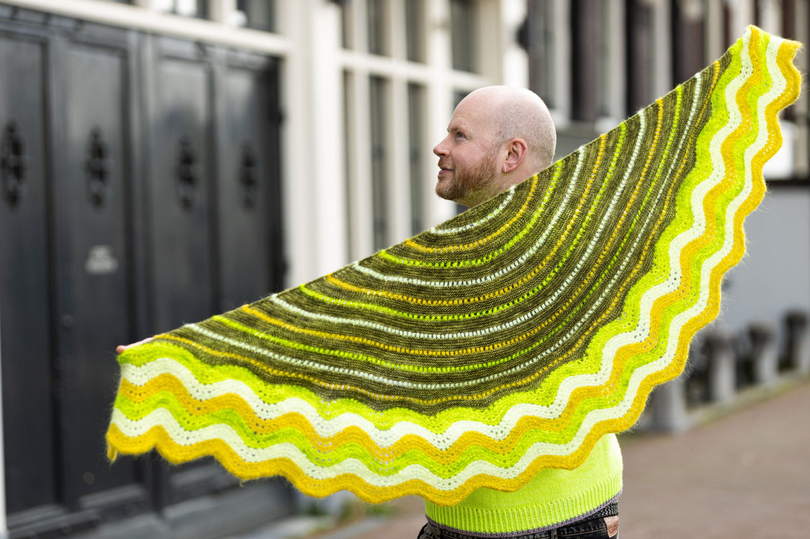 This image shows Stephen West wearing a  beautifully knitted sample of the Suri Shimmy Shawl in green and yellow gradients. The background shows a street in Amsterdam.