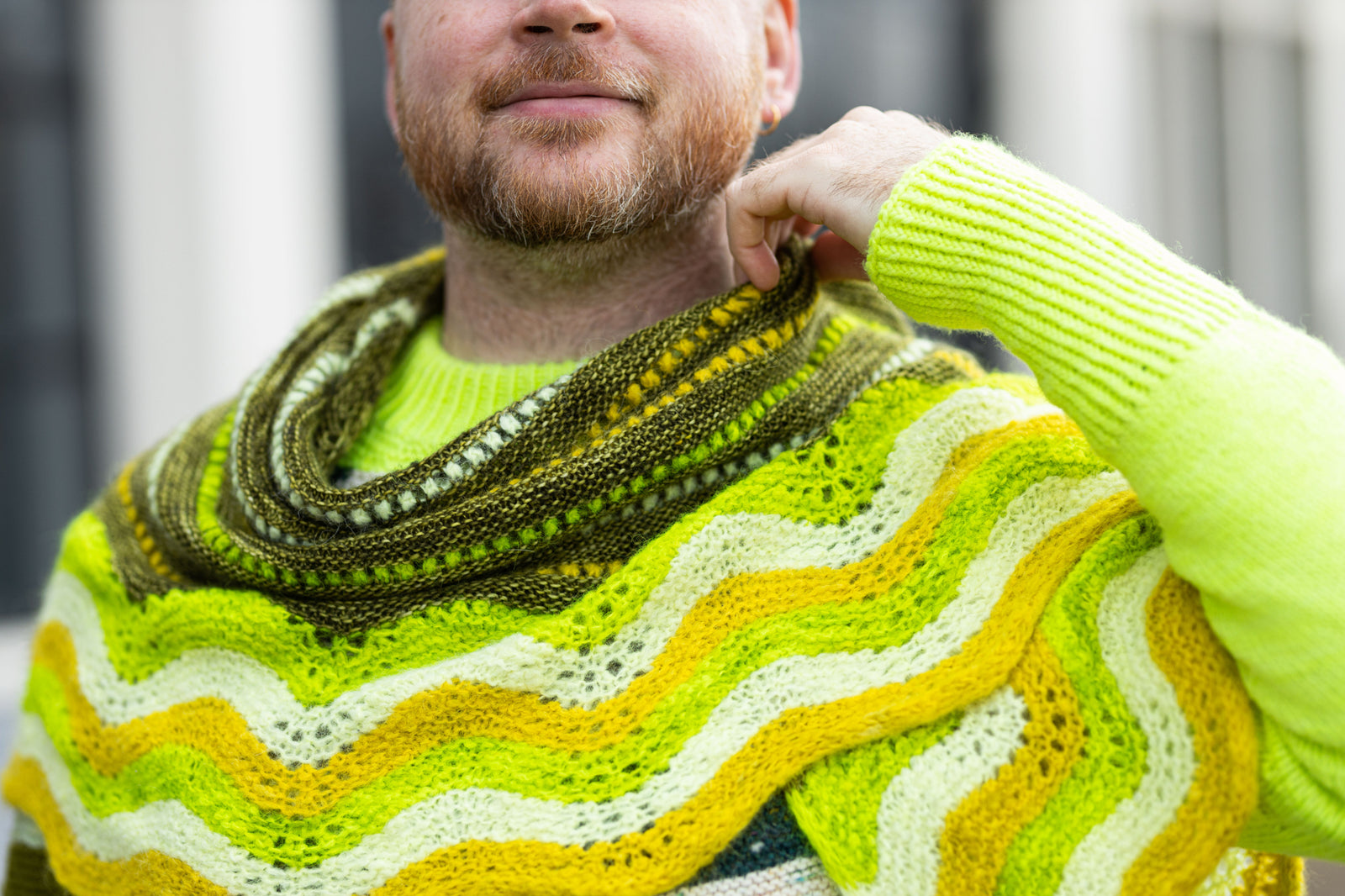 This image shows Stephen West wearing a  beautifully knitted sample of the Suri Shimmy Shawl in green and yellow gradients. 