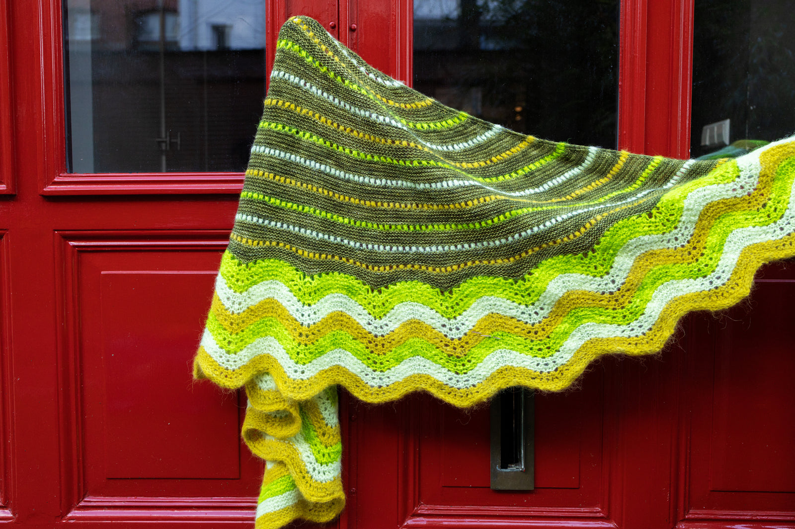 This image shows a  beautifully knitted sample of the Suri Shimmy Shawl in green and yellow gradients hung on a red door. 