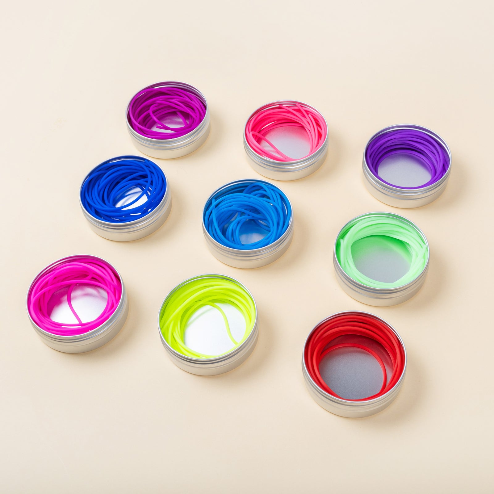 This image shows 9 needle cords of the brand The Knitting Barber. The cords have various colours, such as green, purple, blue, and green and they are inside their metal tins. The cords are laid in a square and they stand on a pastel yellow background. 