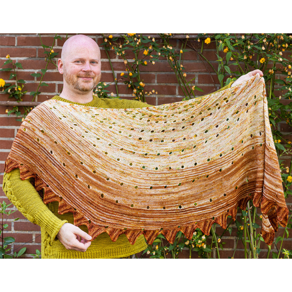 This image shows Stephen West holding a beautifully knitted sample of the Camp Craft Shawl by Westknits, part of the 2025 YAL. The sample is in warm, brown tones. 