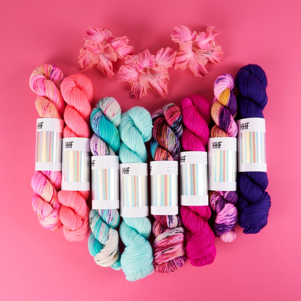 Six skeins of variegated yarn in pink, teal, red, purple, arranged in a heart shape with pink flowers on a solid pink background. Each skein has a white paper label with a colorful stripe pattern and the brand name 'hff' visible.