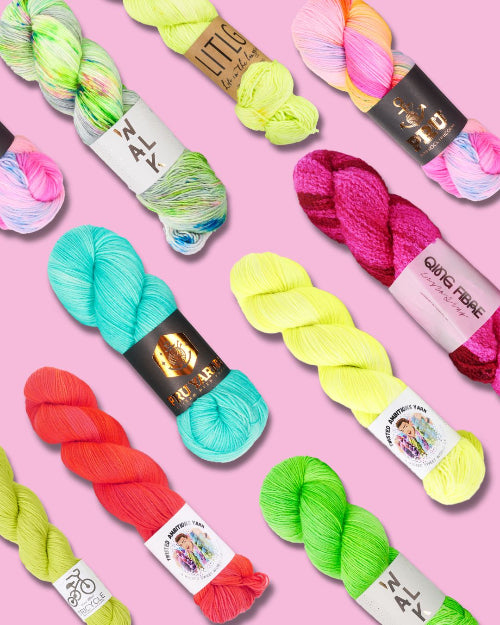 A flat lay of multiple skeins of colorful yarn arranged diagonally on a solid pink background. The yarns come in vibrant shades of neon yellow, turquoise, magenta, coral, lime green, and pastel rainbow blends.
