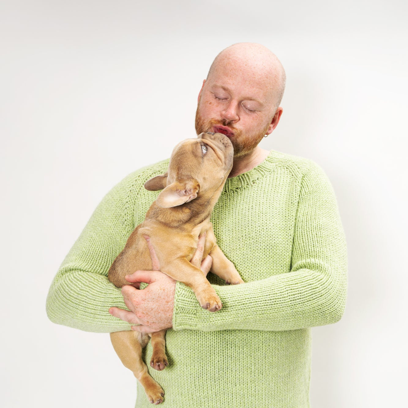 This image shows Stephen West wearing a beautifully knitted sample of the Chunky Raglan Sweater from Westknits. It was knitted with West Wool Cargo. Stephen holds a little puppy in his arms. He stands on a white background. 