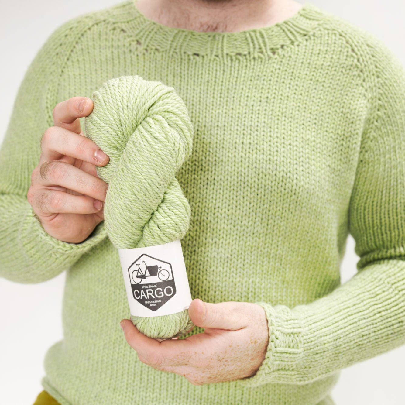 This image shows Stephen West wearing a beautifully knitted sample of the Chunky Raglan Sweater from Westknits. It was knitted with West Wool Cargo. Stephen holds a skein of Cargo in Cucumber. He stands on a white background. 