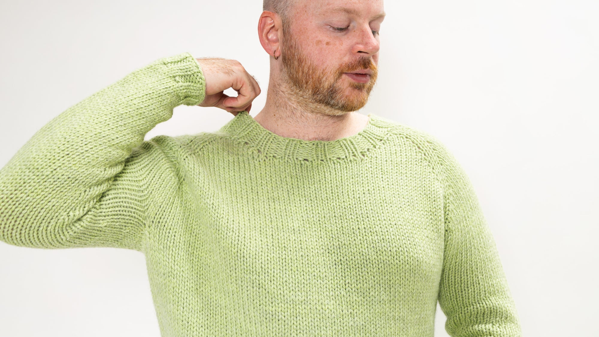 This image shows Stephen West wearing a beautifully knitted sample of the Chunky Raglan Sweater from Westknits. It was knitted with West Wool Cargo. Stephen holds a little puppy in his arms. He stands on a white background. 