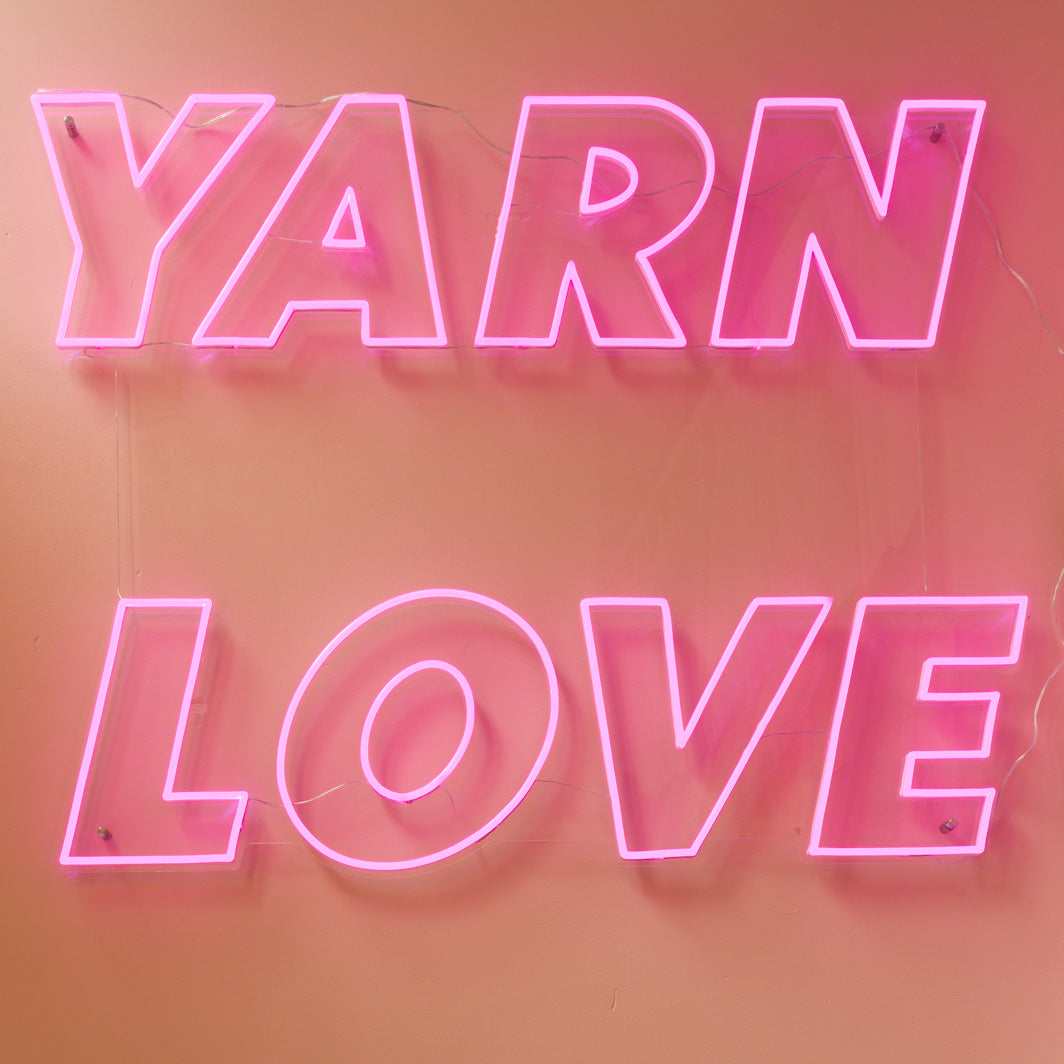 This image shows the text "Yarn Love" in pink neon letters on a pink-orange background. 