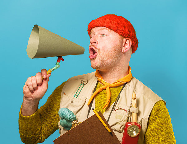 The image shows Stephen West dressed in camping core, using a handmade megaphone. The background is bright blue.  