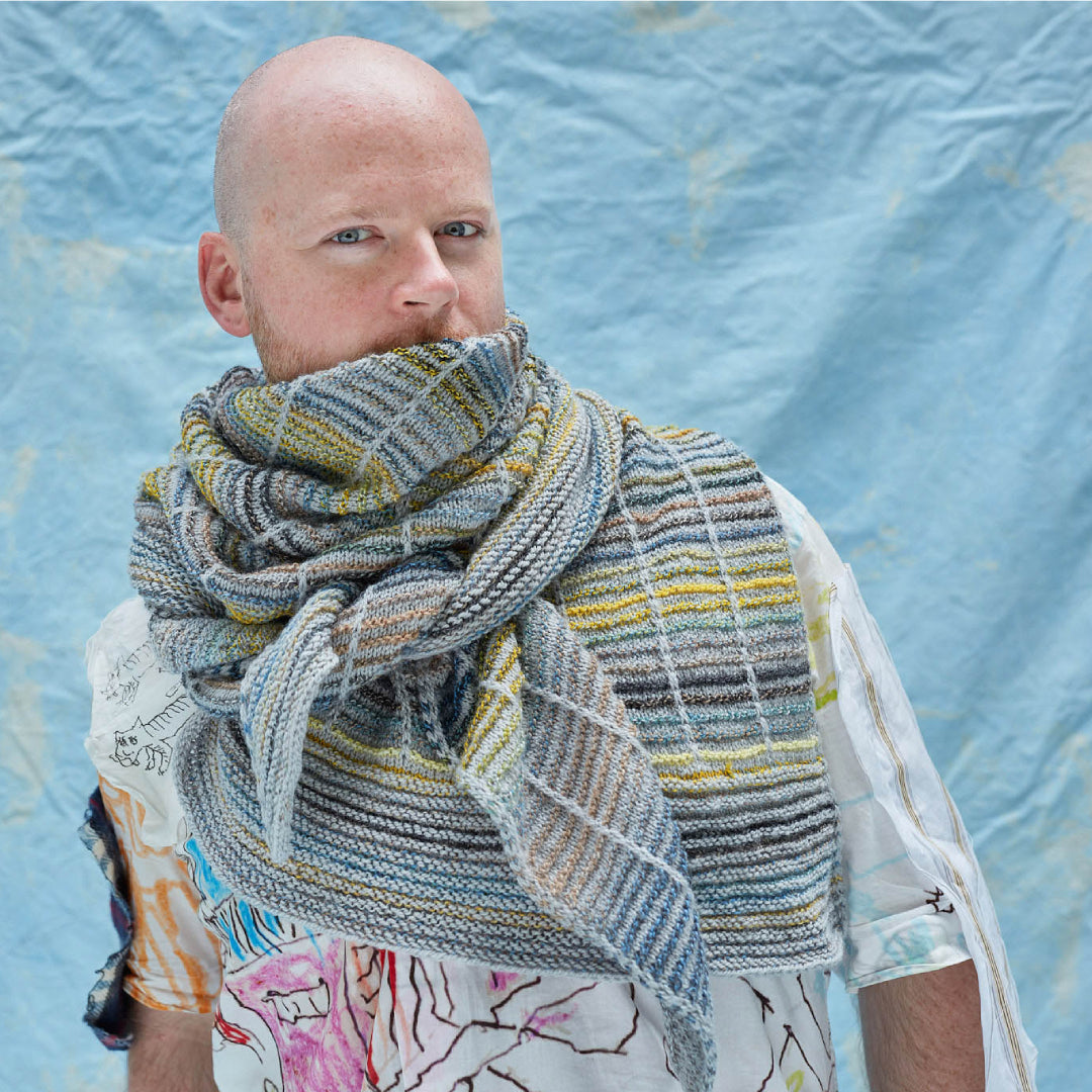 This image shows Stephen West wearing a beautifully knitted sample of the Botanic Shawl by Westknits around his neck. The sample is knitted in yellow, grey, and blue pastel colours. It stands on a light blue background. 
