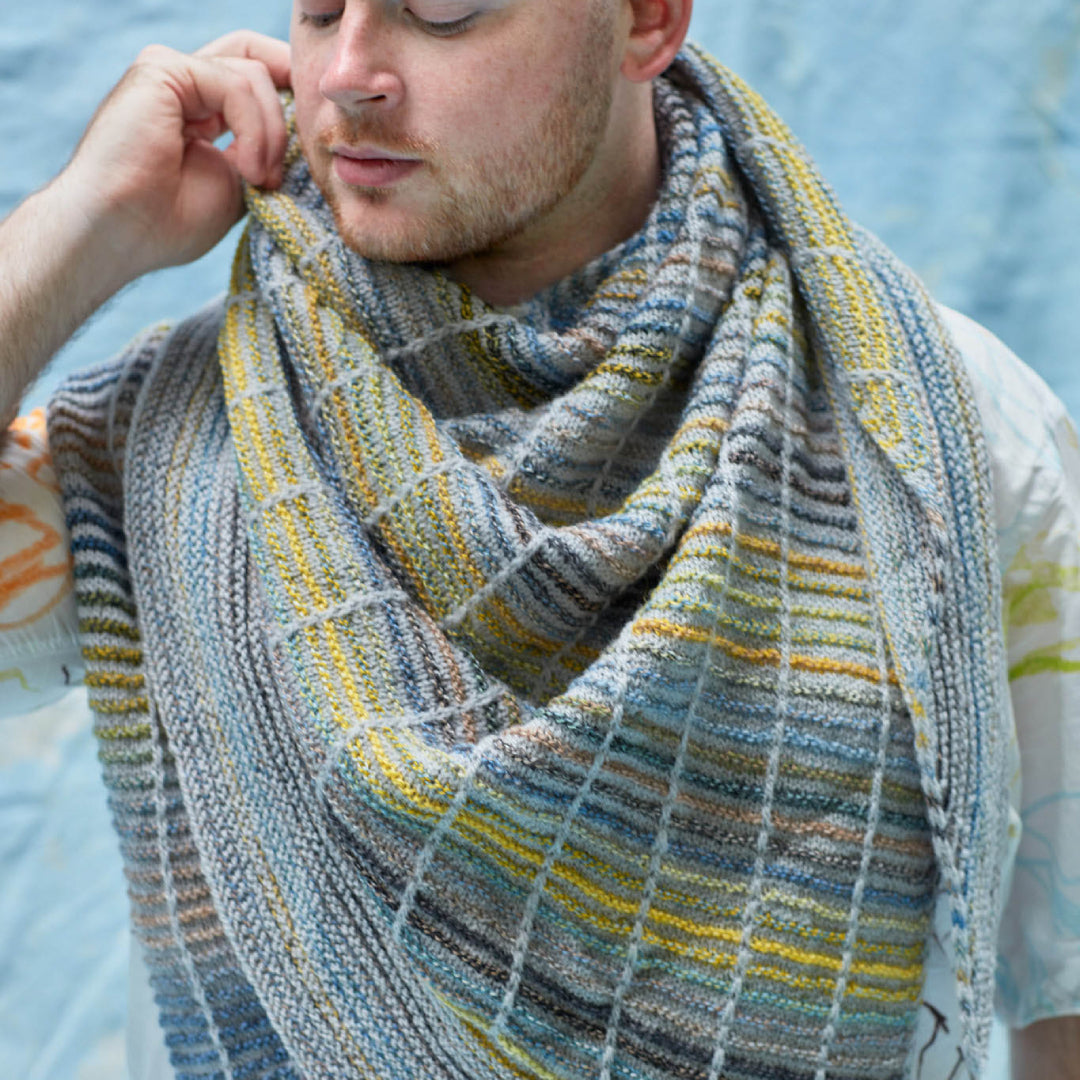 This image shows Stephen West wearing a beautifully knitted sample of the Botanic Shawl by Westknits around his neck. The sample is knitted in yellow, grey, and blue pastel colours. It stands on a light blue background. 