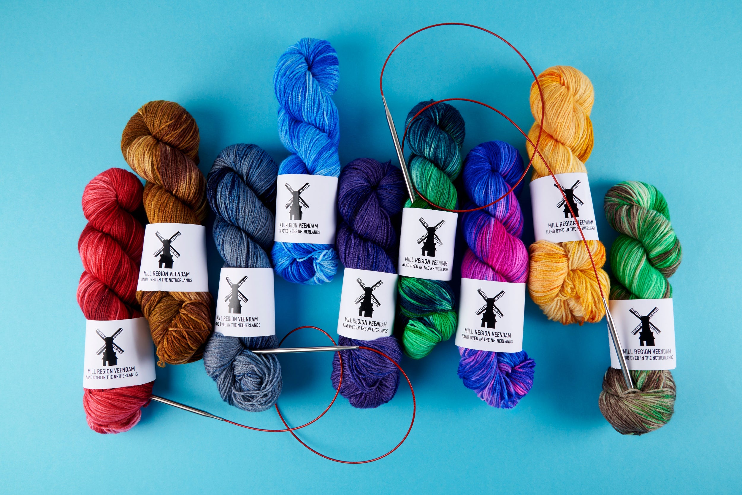 A collection of colorful knitting yarn skeins with circular needles, arranged on a bright blue background. Each skein has a white paper label with a black windmill logo