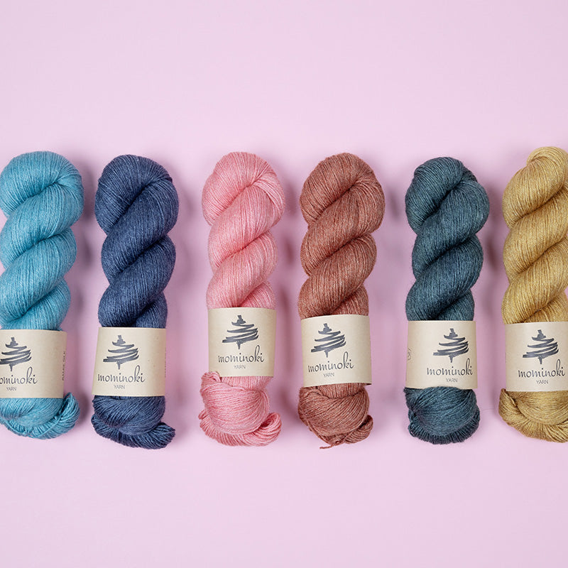 This image shows some skeins of Mominoki Yarn in various colours on a pink background. 