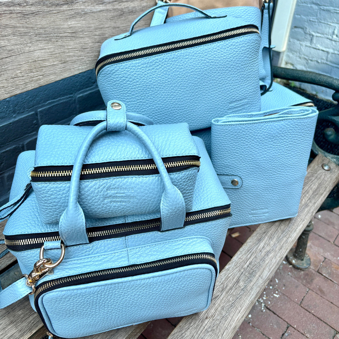A coordinated set of Icy Blue pebbled leather handbags from Re:Designed and Westknits arranged on a wooden bench outdoors.