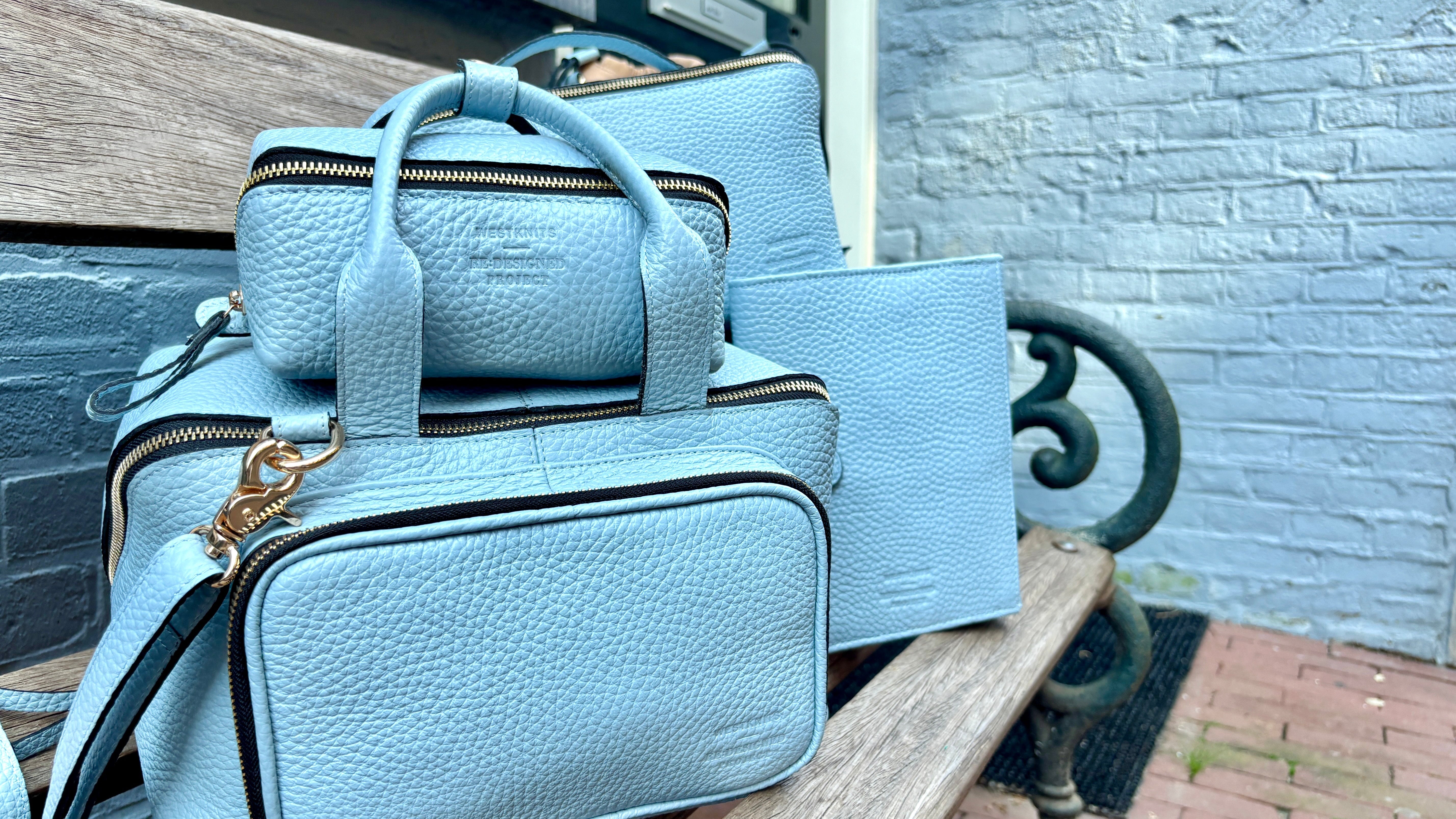 A light blue pebbled leather handbag set with matching pouches and a tote bag, displayed on a wooden bench outdoors next to a gray brick wall.