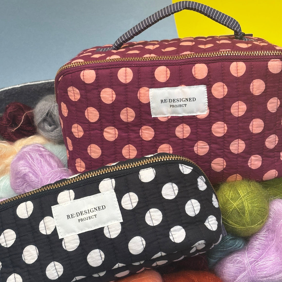 Two quilted zippered cosmetic bags from Redesigned Project, one burgundy with pink polka dots and one black with white polka dots, displayed in a tray filled with colorful balls of yarn.