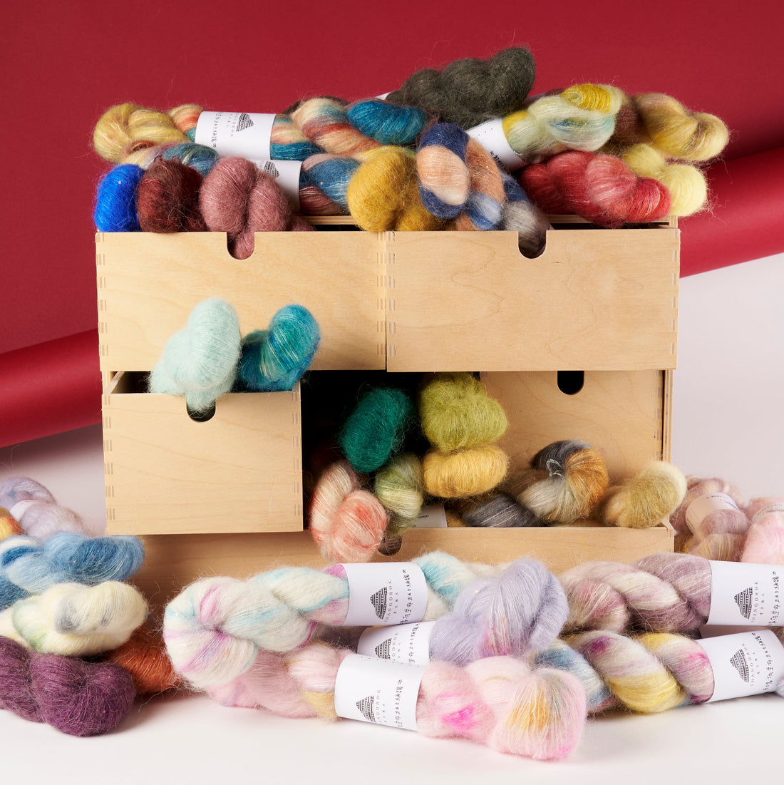 A wooden box filled with colorful variegated wool yarn skeins, with additional skeins spilled in front on a white surface against a red backdrop.