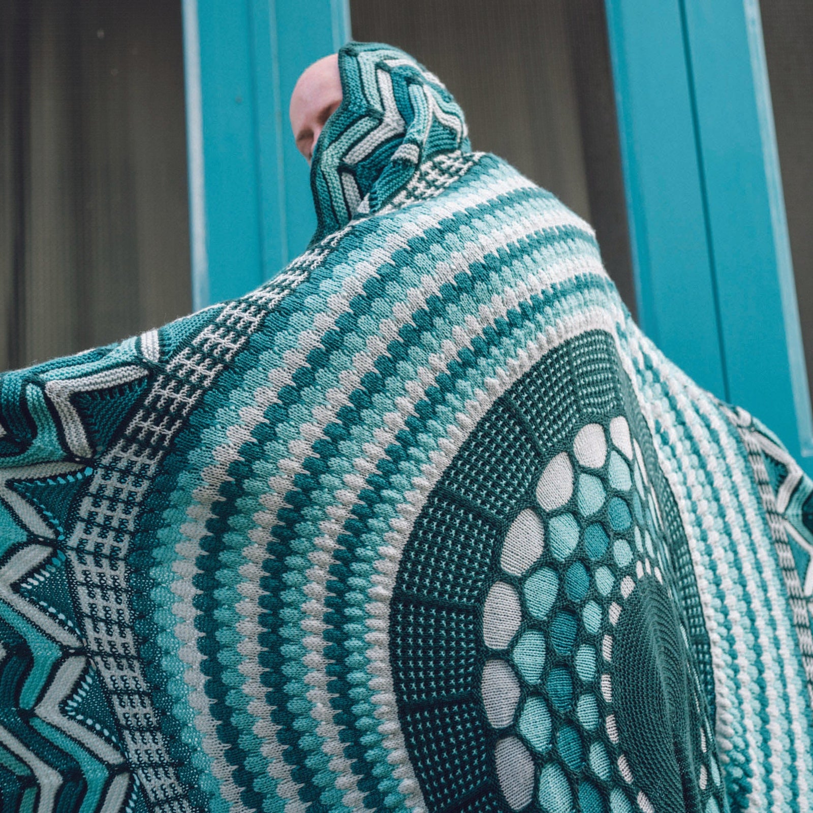This image shows Stephen West wearing a beautifully knitted sample of the Slipstravaganza Blanket in cool blue and turquoise tones. 