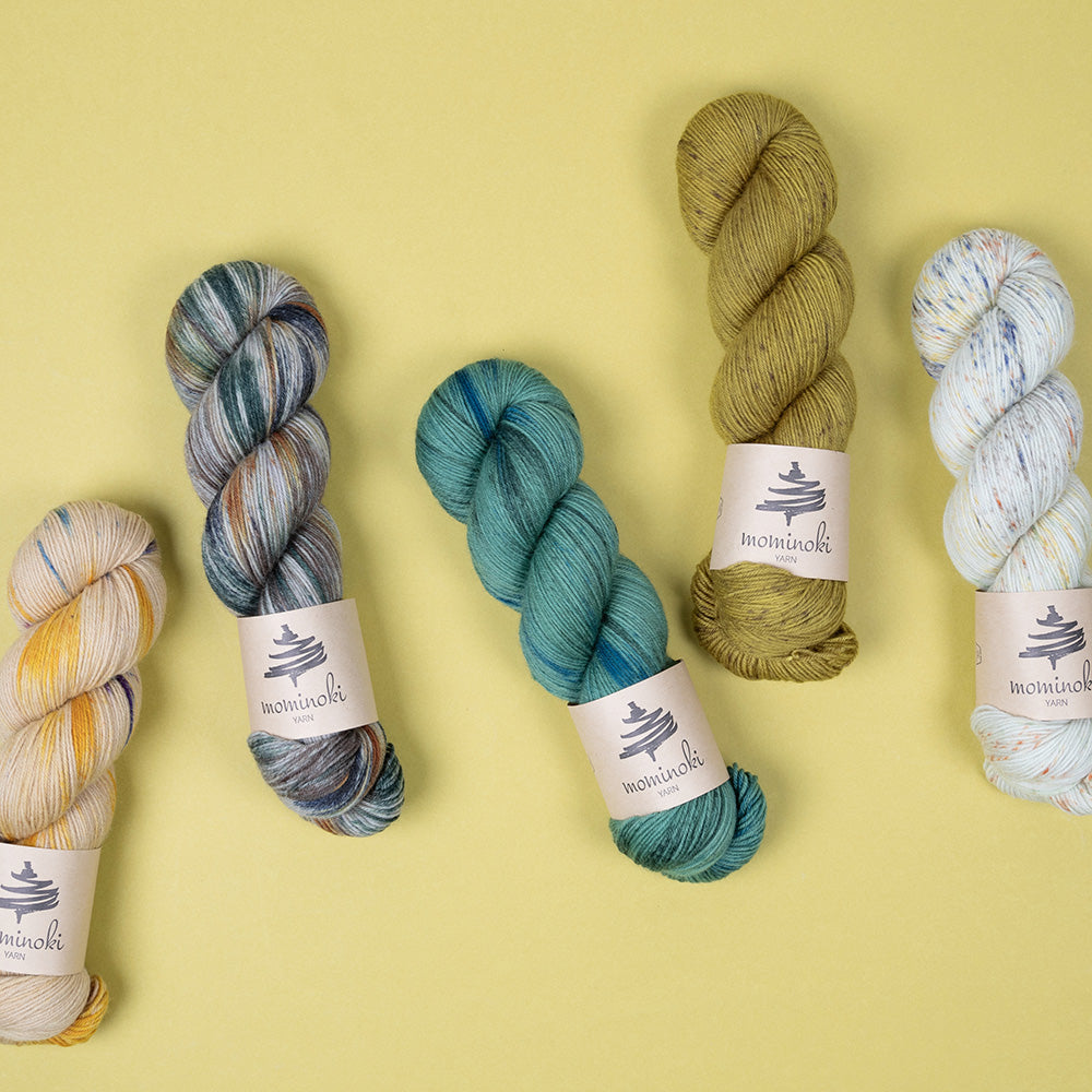 This image shows 5 skeins of Mominoki Yarn in various shades of green on a light green background. 