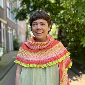 This image shows designer Malia Joseph wearing a beautifully knitted shawl in warm, poppy tones, including yellow and pink. The background shows a street with trees and houses on each side. The shawl is named "Stitchscape Shawl" and it was originally designed for the 2024 Yarn Along. 